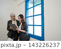 Two businesswomen collaborating with tablet beside bright blue window in modern office, smiling during workplace review, professional teamwork, planning, communication, and urban corporate setting 143226237