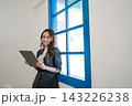 Young professional woman standing beside bright blue window frame holding tablet in modern office interior with soft daylight, smiling confidently, wearing tailored business suit and white blouse 143226238