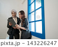Two professional women sharing tablet beside bright blue window in modern office, smiling during collaborative work, planning, review, and friendly business discussion indoors 143226247