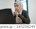 Thoughtful young woman in hijab working at laptop with focused expression in bright office interior, hands clasped near face, modern professional portrait with soft natural light 143226249