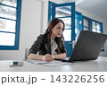 Focused young businesswoman writing beside laptop in bright modern office, working through documents with concentrated expression, blue framed windows, clean workspace, professional daytime setting 143226256