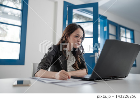 Focused young businesswoman writing beside laptop in bright modern office, working through documents with concentrated expression, blue framed windows, clean workspace, professional daytime setting Focused young businesswoman writing beside laptop in bright modern office, working through documents with concentrated expression, blue framed windows, clean workspace, professional daytime setting 143226256