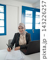 Smiling woman in hijab writing notes at desk beside laptop in bright office with blue windows, professional portrait showing focused work, education, planning, and modern business environment 143226257