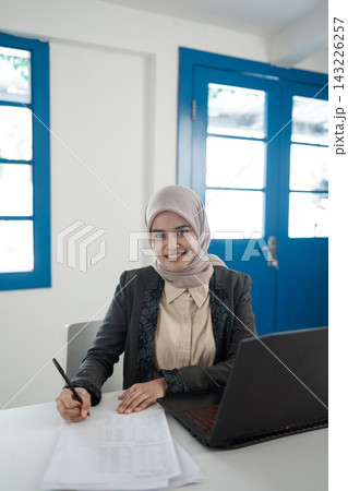 Smiling woman in hijab writing notes at desk beside laptop in bright office with blue windows, professional portrait showing focused work, education, planning, and modern business environment Smiling woman in hijab writing notes at desk beside laptop in bright office with blue windows, professional portrait showing focused work, education, planning, and modern business environment 143226257