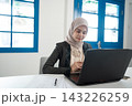 Focused young woman in hijab working on laptop at bright modern office desk with blue windows, papers, notebook, and soft daylight, showing professional online business productivity 143226259