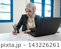 Young Muslim businesswoman in hijab writing on paper beside laptop at bright office desk, focused on paperwork, planning, and administrative work in modern workspace 143226261
