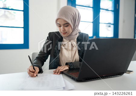 Young Muslim businesswoman in hijab writing on paper beside laptop at bright office desk, focused on paperwork, planning, and administrative work in modern workspace Young Muslim businesswoman in hijab writing on paper beside laptop at bright office desk, focused on paperwork, planning, and administrative work in modern workspace 143226261