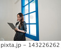 Smiling businesswoman using digital tablet beside bright blue window in modern office interior with clean white wall, elegant suit, soft daylight, professional workspace, calm corporate atmosphere 143226262
