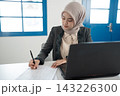 Muslim businesswoman in hijab writing notes at office desk beside laptop under bright window light, focused on paperwork, planning, administration, and professional tasks 143226300