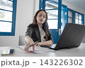 Young businesswoman in modern office reaching across desk beside open laptop and papers, focused on work in bright interior with blue doors and windows 143226302