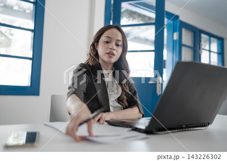 Young businesswoman in modern office reaching across desk beside open laptop and papers, focused on work in bright interior with blue doors and windows Young businesswoman in modern office reaching across desk beside open laptop and papers, focused on work in bright interior with blue doors and windows 143226302
