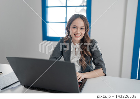 Smiling businesswoman working at laptop in bright office with window light, wearing blazer and white blouse, seated at desk with documents, showing confident professional attitude Smiling businesswoman working at laptop in bright office with window light, wearing blazer and white blouse, seated at desk with documents, showing confident professional attitude 143226338