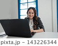 Smiling young businesswoman working on laptop at bright modern office desk with blue window frame, focused posture, professional outfit, natural daylight, remote work concept 143226344
