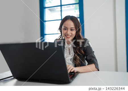 Smiling young businesswoman working on laptop at bright modern office desk with blue window frame, focused posture, professional outfit, natural daylight, remote work concept Smiling young businesswoman working on laptop at bright modern office desk with blue window frame, focused posture, professional outfit, natural daylight, remote work concept 143226344
