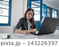 Focused young businesswoman writing notes beside laptop in bright modern office with blue windows, concentrating on paperwork, planning daily tasks, and digital work 143226397