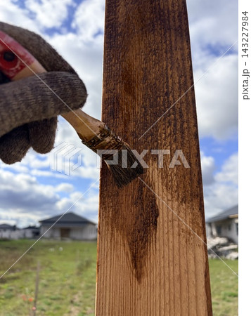 A worker applies dark wood stain with a brush to the wood outdoors. A worker applies dark wood stain with a brush to the wood outdoors. 143227984