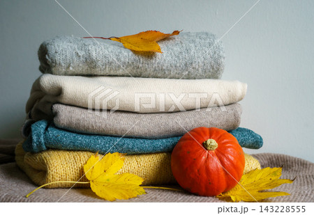 An orange pumpkin and autumn leaves rest beside folded knitwear. Concept of autumn, warmth, harvest, comfort, clothing, seasonality and home comfort 143228555