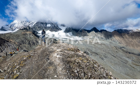 View of Grossglockner from near Studl Hut in the Austrian Alps, dramatic glaciated mountain landscape with hikers on a rocky ridge beneath clouds and blue sky 143230372