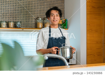 キッチンで鍋を持ち笑顔を見せるエプロン姿の男性。料理や家事を楽しむ暮らしのイメージ キッチンで鍋を持ち笑顔を見せるエプロン姿の男性。料理や家事を楽しむ暮らしのイメージ 143230866