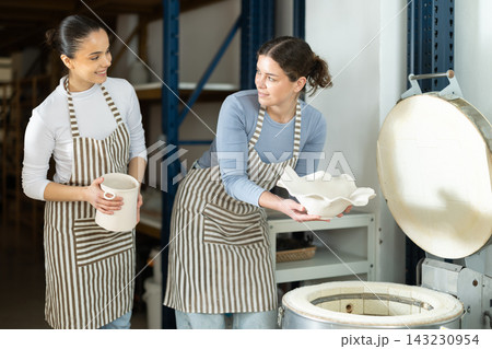 Two female potters take finished ceramic dishes, various mugs and a vase out of oven Two female potters take finished ceramic dishes, various mugs and a vase out of oven 143230954