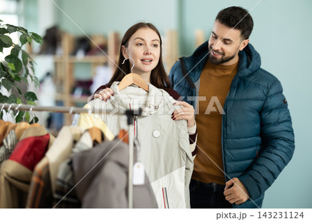 Happy couple examine and selecting clothes for cold season in modern store, man trying new coat 143231124