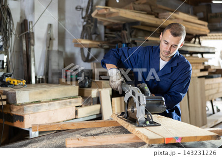 Male employee of workshop cuts wooden board into narrow parts using circular saw 143231226