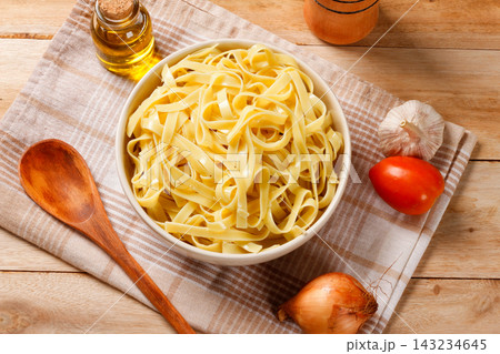 Cooked fresh fettuccine served in a bowl on a cloth napkin and wooden table, with olive oil, garlic, tomato, and onion. 143234645