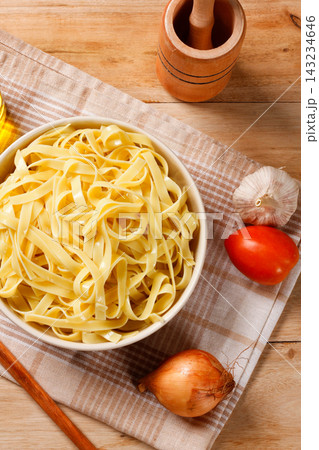 Cooked fettuccine-style pasta served in a bowl on a rustic wooden surface, with a cloth and Mediterranean culinary ingredients. 143234646