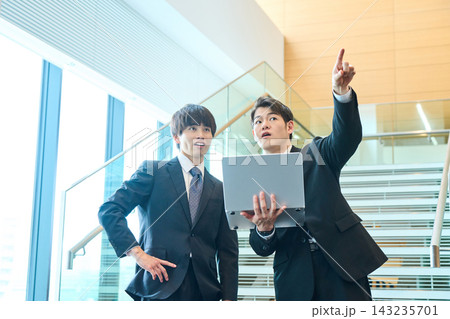 オフィスの階段前でPCを手に上を指差し打ち合わせをするスーツ姿の男性社員2人 143235701