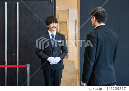 イベント会場の入り口で笑顔でお客様を出迎えるスーツ姿の受付案内スタッフ イベント会場の入り口で笑顔でお客様を出迎えるスーツ姿の受付案内スタッフ 143239237