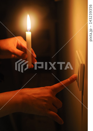 A man holds a candle while reaching for a light switch in a dark room. electricity crisis concept. A man holds a candle while reaching for a light switch in a dark room. electricity crisis concept. 143239356