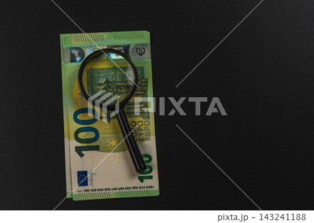 Examining a euro banknote with a magnifying glass on a dark surface 143241188