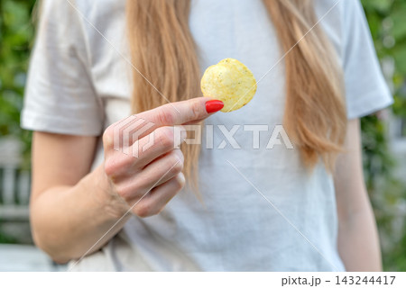 Woman's hand with red nail polish holding up a single golden potato chip, suggesting an indulgent snack or craving for crunchy fast food in a casual outdoor setting Woman's hand with red nail polish holding up a single golden potato chip, suggesting an indulgent snack or craving for crunchy fast food in a casual outdoor setting 143244417