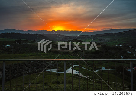 Bright orange sunset over mountains and valley seen through a railing in Ronda Bright orange sunset over mountains and valley seen through a railing in Ronda 143246675