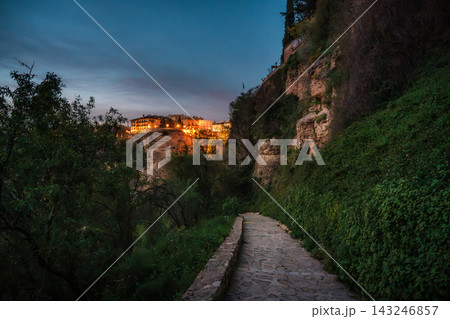 Stone footpath below Ronda cliffs toward illuminated clifftop buildings at dusk Stone footpath below Ronda cliffs toward illuminated clifftop buildings at dusk 143246857