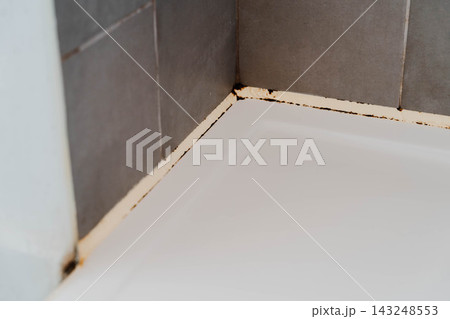 Black mold and rust stains spread along the aging silicone sealant seam where a white bathtub meets grey wall tiles. Detail for bathroom maintenance, waterproofing repair and mildew removal. Black mold and rust stains spread along the aging silicone sealant seam where a white bathtub meets grey wall tiles. Detail for bathroom maintenance, waterproofing repair and mildew removal. 143248553