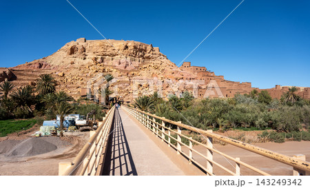 Visitors walk across a bridge leading to Ait Benhaddou. This site showcases traditional clay architecture set against a desert backdrop. The area holds rich cultural significance and history. 143249342