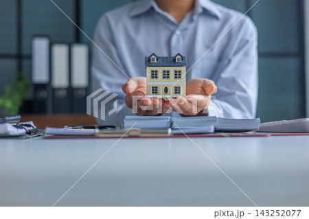 Businessman, Banker, Realtor at office desk holding a house model with money, calculator, and financial documents or contract on the table 143252077