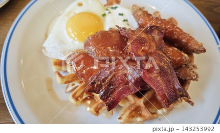 Breakfast plate featuring golden waffles topped with crispy bacon, fried egg, and sausage links, served on a white plate with blue rim 143253992