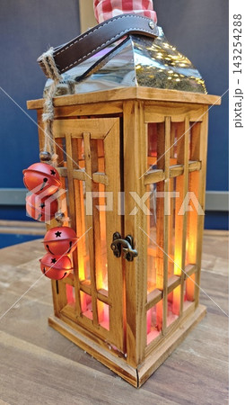 Wooden lantern with glass panels and red bells, illuminated from within, placed on a wooden surface against a blue background in a cozy indoor setting 143254288