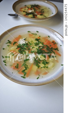 Bowl of clear soup with meatballs, carrots, potatoes, and herbs on a white table, another bowl of soup slightly blurred in the background Bowl of clear soup with meatballs, carrots, potatoes, and herbs on a white table, another bowl of soup slightly blurred in the background 143254475