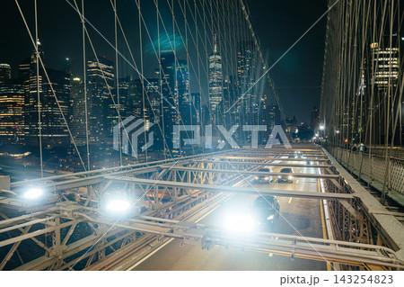 Vehicles moving along the Brooklyn Bridge roadway with suspension cables and bright city lights at night. Lower Manhattan skyline with illuminated skyscrapers and One World Trade Center visible in the Vehicles moving along the Brooklyn Bridge roadway with suspension cables and bright city lights at night. Lower Manhattan skyline with illuminated skyscrapers and One World Trade Center visible in the 143254823