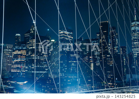 Illuminated skyscrapers of Lower Manhattan seen through the suspension cables of the Brooklyn Bridge at night. Urban architecture scene with glowing city lights and dramatic bridge structure. 143254825