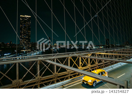 Yellow taxi driving across the Brooklyn Bridge with Manhattan skyline and illuminated buildings in New York City at night. Urban traffic scene with bridge cables, city lights, and reflections over the 143254827