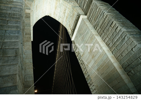 Low angle view of the historic Brooklyn Bridge stone arch and suspension cables against the dark night sky in New York City. Dramatic architectural perspective highlighting the massive stone structure 143254829