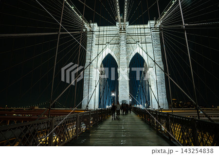 People walk along the wooden pedestrian walkway of the Brooklyn Bridge at night in New York City. The illuminated bridge tower and suspension cables create a dramatic perspective of the historic People walk along the wooden pedestrian walkway of the Brooklyn Bridge at night in New York City. The illuminated bridge tower and suspension cables create a dramatic perspective of the historic 143254838