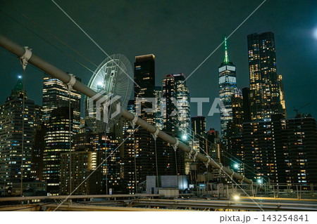 Tall skyscrapers of Manhattan glow with lights at night as seen from the Brooklyn Bridge in New York City. The dense city skyline highlights the financial and business center of the United States. 143254841