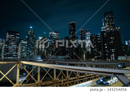 Illuminated skyscrapers of Lower Manhattan rise behind the steel roadway of the Brooklyn Bridge at night. Bright city lights and modern architecture create a dramatic New York City skyline. Illuminated skyscrapers of Lower Manhattan rise behind the steel roadway of the Brooklyn Bridge at night. Bright city lights and modern architecture create a dramatic New York City skyline. 143254856
