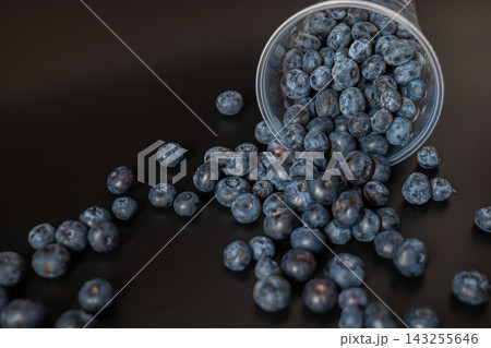An overturned plastic cup releases blueberries in a diagonal trail across a dark surface. Fruit motion and abundance express harvest, snack preparation, and food spill concepts. An overturned plastic cup releases blueberries in a diagonal trail across a dark surface. Fruit motion and abundance express harvest, snack preparation, and food spill concepts. 143255646