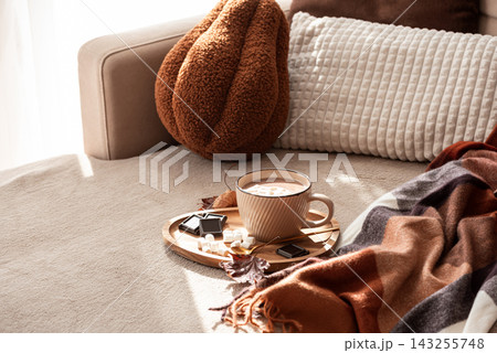 Cozy warm hot chocolate with marshmallows and chocolate squares on a tray next to a blanket and pillows indoors 143255748